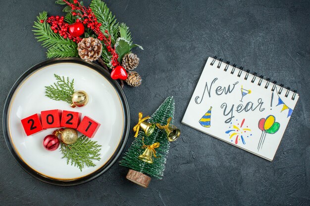 Vue de dessus des accessoires de décoration numéros sur une assiette branches de sapin conifère cône et cahier avec nouvel an écrit et dessin sur fond sombre