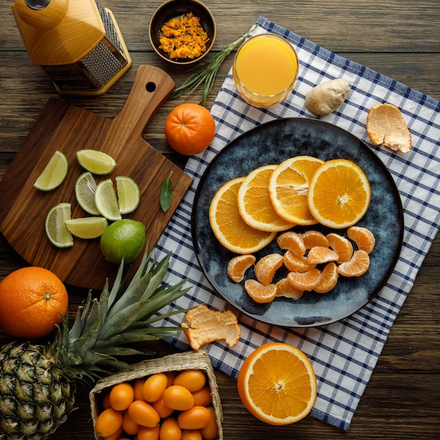Photo gratuite vue de dessus des agrumes sous forme de tranches d'orange et de mandarine dans une assiette avec du jus d'orange gingembre coquille de mandarine sur tissu à carreaux avec zeste d'orange kumquats citron vert ananas avec râpe sur fond en bois