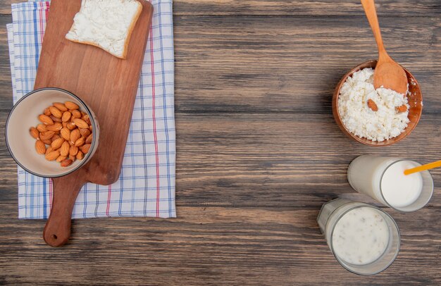 Vue de dessus des amandes dans un bol et une tranche de pain sur une planche à découper sur un tissu à carreaux et une soupe au lait et au yogourt au fromage cottage sur fond de bois avec espace de copie