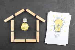 Photo gratuite vue de dessus de l'ampoule idealight dans des blocs de bois en forme de maison ampoule idealight dessin sur le bloc-notes sur fond noir
