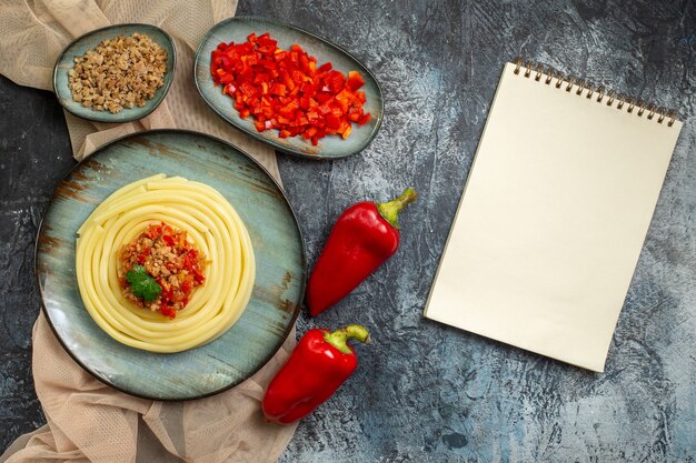 Vue de dessus d'une assiette bleue avec un délicieux repas de pâtes servi avec de la tomate et de la viande sur une serviette de couleur beige hachée et des poivrons entiers et un cahier à spirale