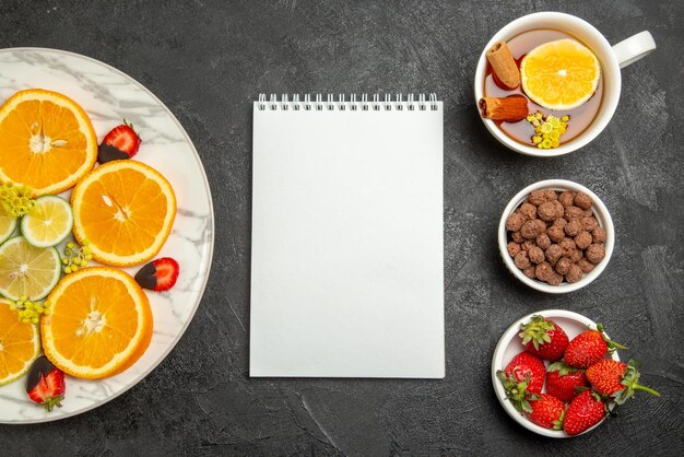 Vue de dessus assiette de fruits assiette de tranches de fraises enrobées de chocolat orange citron à côté du cahier une tasse de thé noisettes et fraises