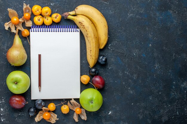 Vue de dessus bananes jaunes avec pommes vertes fraîches poires prunes crayon bloc-notes et cerises douces sur le bureau bleu foncé santé vitamine fruit berry