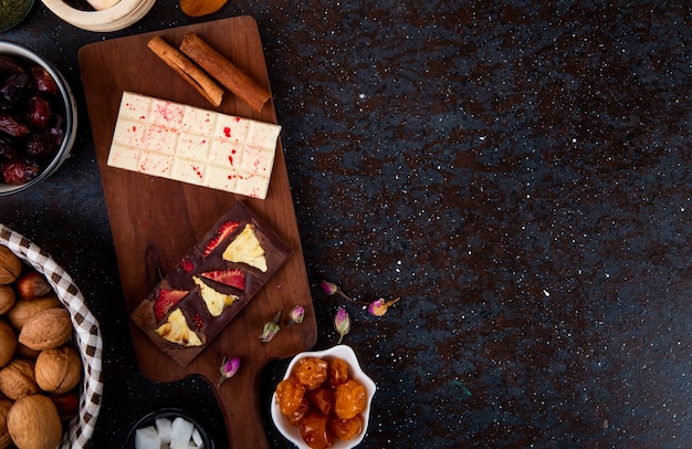 Vue de dessus des barres de chocolat noir et blanc avec des bâtons de cannelle sur planche de bois et avec des fruits secs et des noix sur fond noir avec copie espace