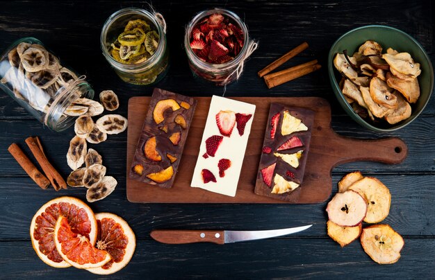 Vue de dessus des barres de chocolat noir et blanc sur une planche à découper en bois avec divers fruits en tranches séchées sur fond noir