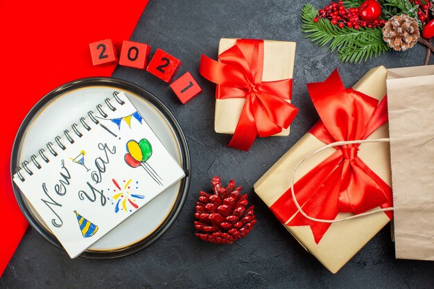 Vue de dessus de beaux cadeaux et cahier avec des dessins de nouvel an sur une plaque de conifères cône de branches de sapin numéros sur un tableau sombre