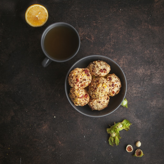 Vue de dessus des biscuits aux pistaches dans un bol avec une tasse de café et de citron alignés en diagonale sur une texture brun foncé.