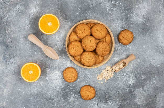 Vue de dessus des biscuits faits maison sur planche de bois et flocons d'avoine avec des oranges sur une surface grise.