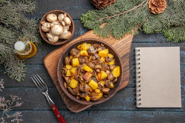 Vue de dessus et bloc-notes pommes de terre et champignons dans un bol sur une planche à découper à côté de la fourchette et du bloc-notes sous un bol d'huile de champignons en bouteille et des branches avec des cônes