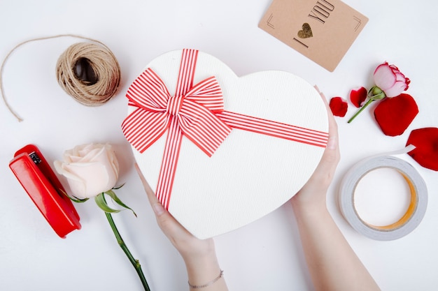 Vue de dessus d'une boîte-cadeau en forme de coeur avec un arc et une rose de couleur blanche et une agrafeuse avec une corde et une carte postale sur fond blanc