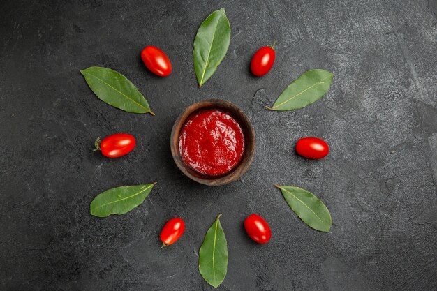 Vue de dessus un bol de ketchup autour des tomates cerises et des feuilles de laurier sur fond sombre