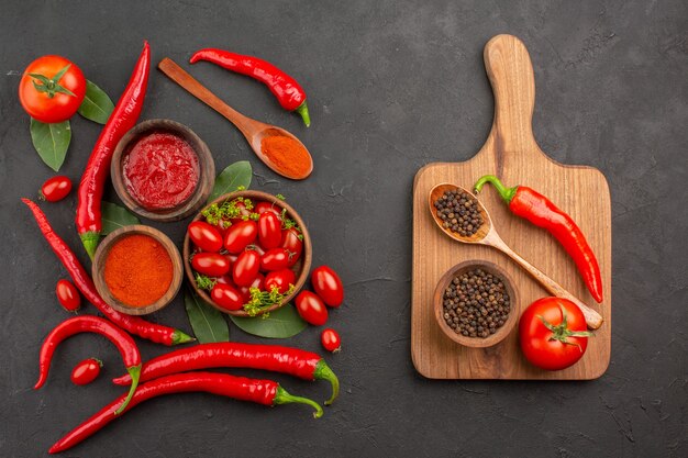 Vue de dessus un bol de tomates cerises piments rouges chauds feuilles de laurier et un bol de poivre noir une cuillère une tomate un poivron rouge sur la planche à découper sur fond noir