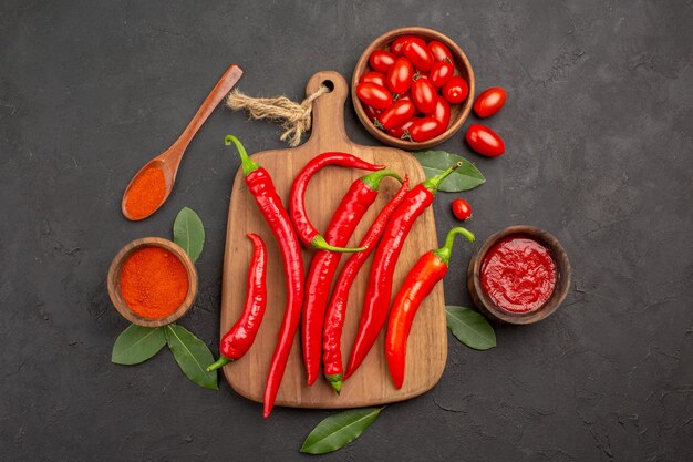 Vue de dessus un bol de tomates cerises piments rouges chauds sur la planche à découper une cuillère en bois feuilles de laurier et bols de ketchup et de poudre de piment sur la table noire