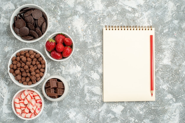 Vue de dessus des bols avec des bonbons fraises chocolats céréales et cacao à gauche et un cahier avec un crayon rouge sur le côté droit de la table gris-blanc