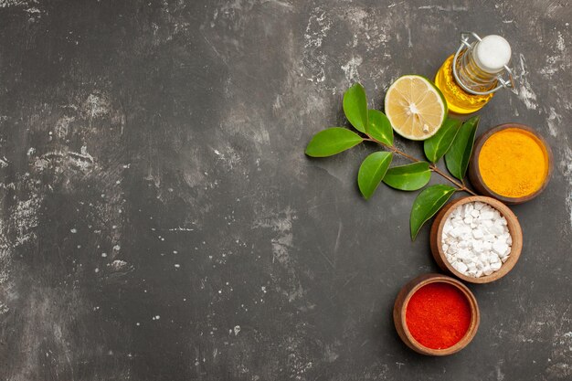 Vue de dessus des bols d'épices d'épices colorées citron avec des feuilles à côté de la bouteille d'huile sur le côté droit de la table sombre