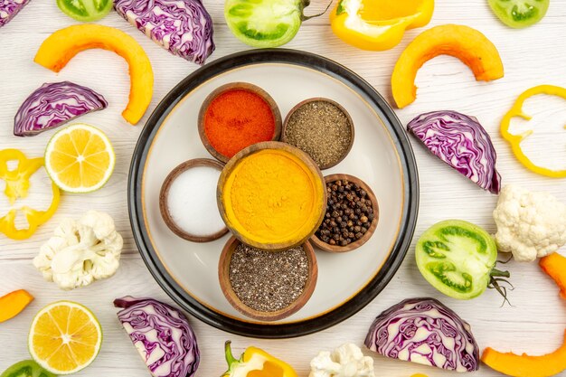 Vue de dessus des bols avec des épices sur plaque ronde sel de curcuma poivre noir poivron rouge légumes coupés sur une surface blanche