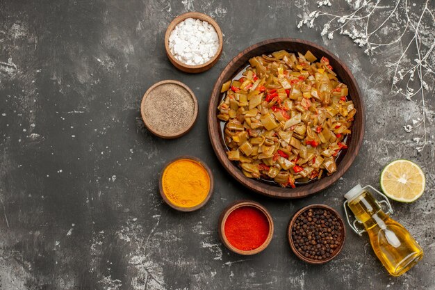 Vue de dessus des bols de nourriture appétissants d'épices colorées bouteille d'huile et de citron autour d'une assiette brune de haricots verts et de tomates à côté des branches d'arbres