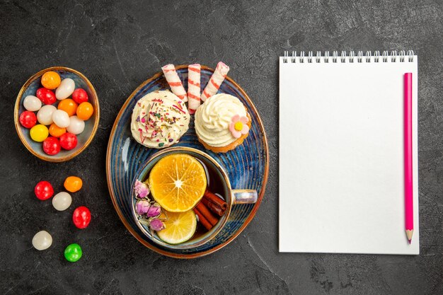 Vue de dessus des bonbons sur l'assiette assiette des cupcakes appétissants bols de bonbons colorés à côté du cahier blanc et du crayon rose sur la table