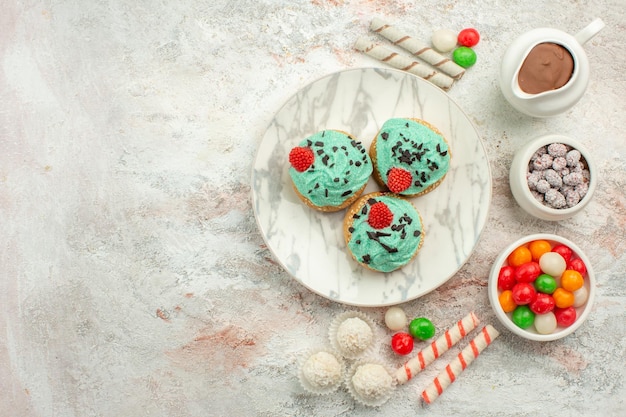 Vue de dessus des bonbons colorés avec des gâteaux crémeux sur un gâteau au thé aux biscuits arc-en-ciel de couleur de surface blanche