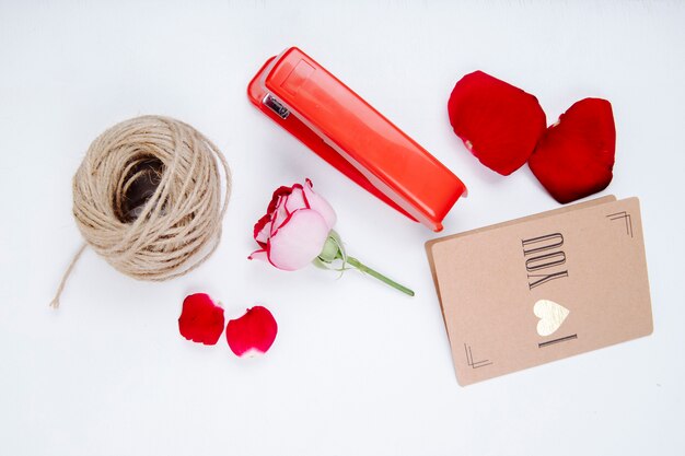 Photo gratuite vue de dessus d'une boule de corde pétales de rose rouges et fleur rose avec petite carte postale et agrafeuse sur fond blanc
