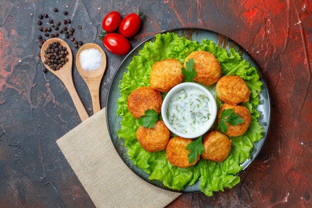 Vue de dessus des boules de fromage sur une assiette avec de la laitue et de la sauce tomates cerises cuillères en bois poivrons noirs sur une surface sombre