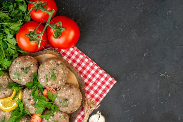 Vue de dessus des boulettes de viande non cuites sur une planche à découper en bois et des légumes frais verts sur le côté droit sur fond sombre