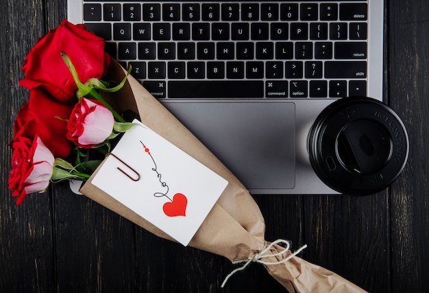 Vue De Dessus Un Bouquet De Roses Rouges En Papier Kraft Avec Carte Postale Attachée Allongé Sur Un Ordinateur Portable Avec Une Tasse De Café En Papier Sur Fond De Bois Foncé