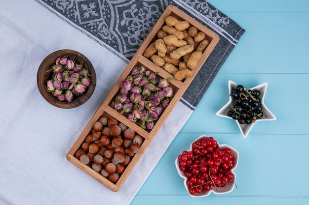 Vue de dessus des boutons de roses séchées aux noisettes et arachides sur une serviette blanche avec des groseilles rouges et noires sur une surface bleue