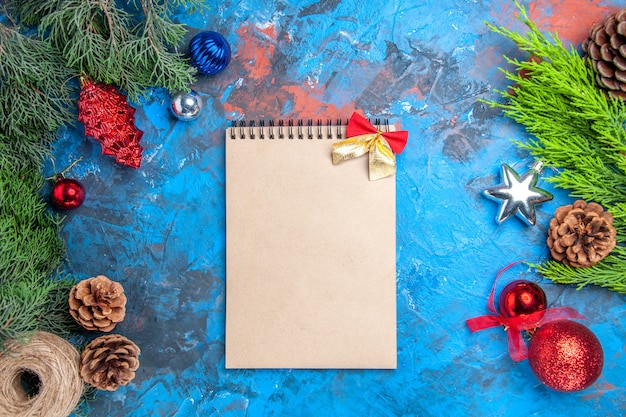 Vue de dessus des branches de pin avec des pommes de pin et des jouets d'arbre de Noël colorés paille enfiler un cahier sur fond bleu-rouge