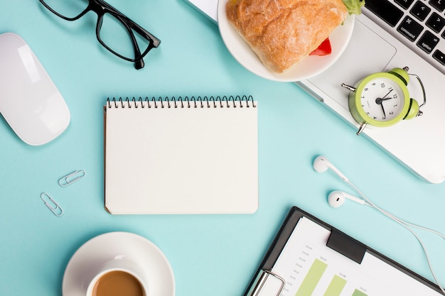 Vue de dessus d'un bureau avec papeterie, ordinateur portable, souris et réveil sur fond bleu