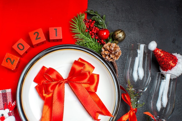 Vue de dessus de cadeau avec des assiettes à dîner ruban rouge accessoires de décoration branches de sapin chaussette de Noël gobelets en verre chapeau de père Noël sur table sombre