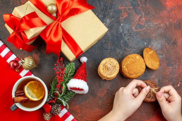 Vue de dessus des cadeaux de noël attachés avec des biscuits au ruban rouge santa hat dans une tasse de thé à la main féminine sur une table rouge foncé