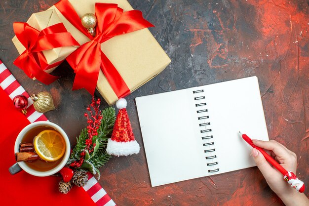 Vue de dessus des cadeaux de Noël attachés avec un ruban rouge stylo bloc-notes de branche d'arbre de Noël dans une main féminine sur une table rouge foncé