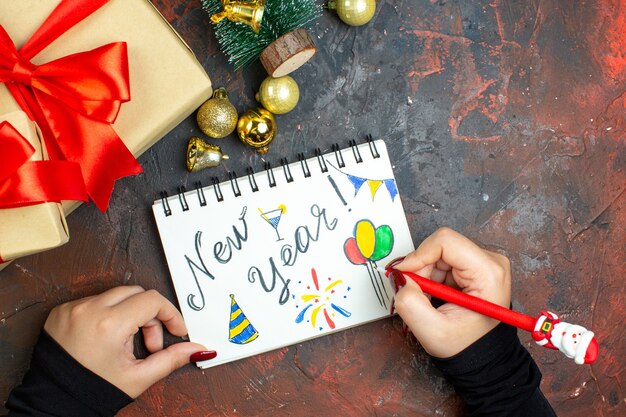 Vue de dessus cadeaux de noël boules de noël dorées mini arbre de noël nouvel an écrit sur un cahier et un stylo dans les mains de la femme sur une table rouge foncé