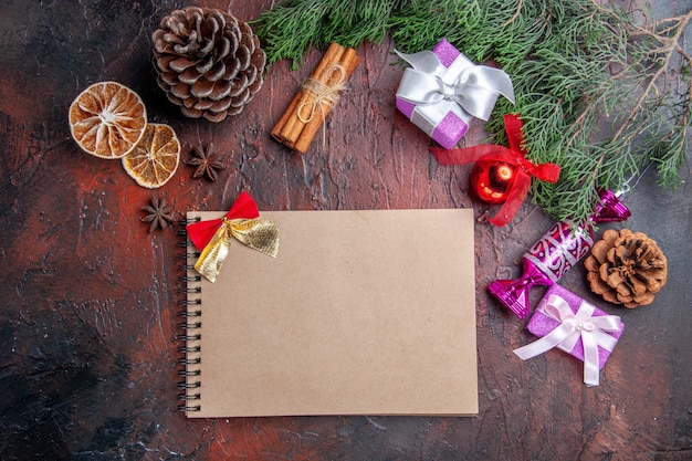 Vue de dessus cadeaux de noël branches de pin avec cône jouets de noël tranches de citron séchées à la cannelle anis un cahier sur une surface rouge foncé