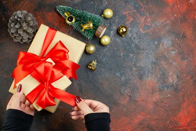 Vue de dessus des cadeaux de noël dans les mains des femmes boules de noël dorées mini arbre de noël sur l'espace de copie de table rouge foncé