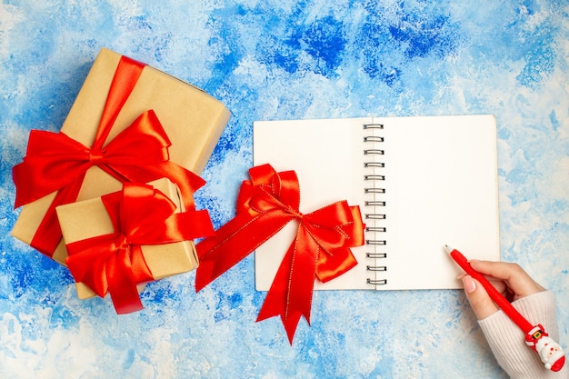 Vue de dessus des cadeaux de Noël de différentes tailles à égalité avec un arc rouge sur un stylo bloc-notes dans une main féminine sur une table bleue