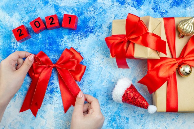 Vue de dessus des cadeaux de vacances petit bonnet de noel blocs de bois arc rouge dans la main féminine sur table bleu blanc