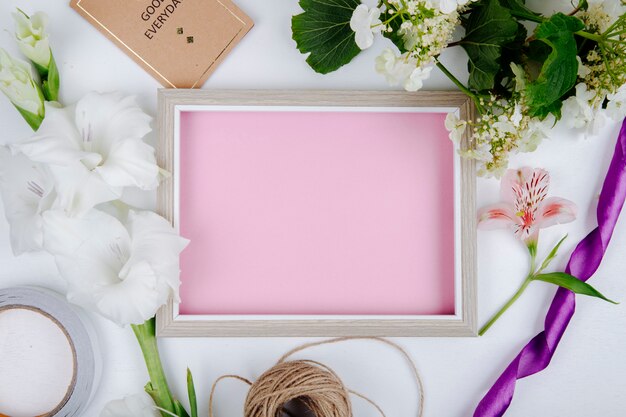 Vue de dessus d'un cadre photo avec une feuille de papier rose petite carte postale corde et fleur de glaïeul de couleur blanche et une branche de viburnum en fleurs sur fond blanc