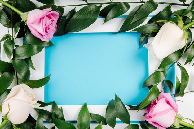 Vue de dessus d'un cadre photo vide avec des roses blanches et roses et ruscus sur fond bleu avec copie espace