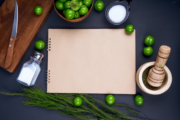 Vue de dessus d'un carnet de croquis, salière, menthe poivrée séchée dans un mortier et prunes vertes aigres dans un bol en bois sur tableau noir