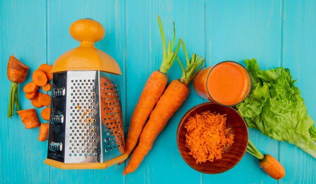 Vue de dessus de carottes entières râpées coupées entières avec de la laitue râpe en métal et du jus de carotte sur la table bleue