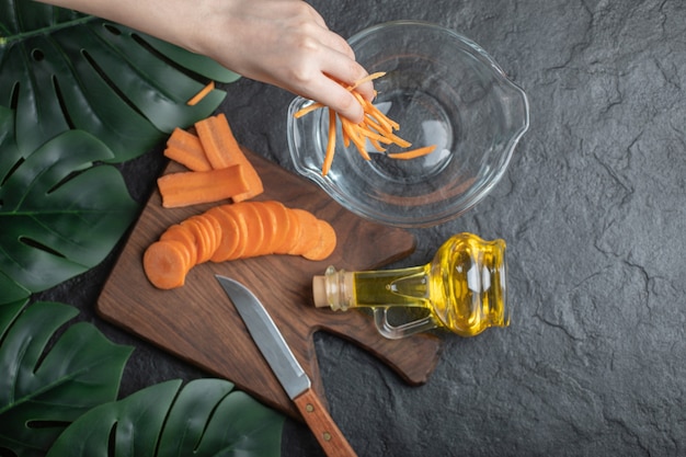 Vue de dessus des carottes tranchées sur une planche à découper en bois et une main féminine met des tranches de carottes dans un bol.