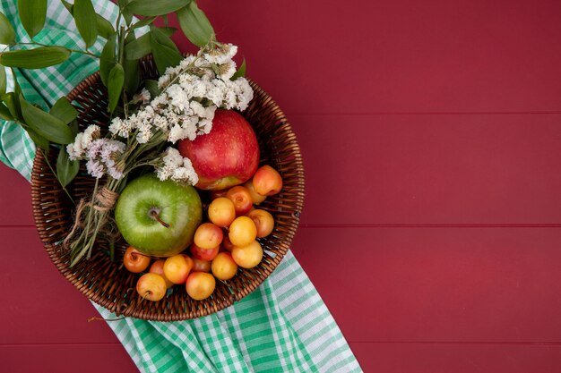 Vue de dessus de la cerise blanche avec des pommes et des fleurs colorées dans un panier sur une surface rouge