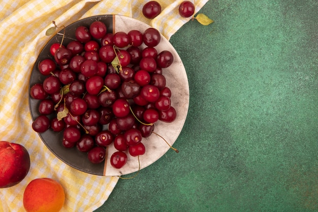 Vue de dessus des cerises en assiette avec pêche et abricot sur tissu à carreaux sur fond vert avec espace copie