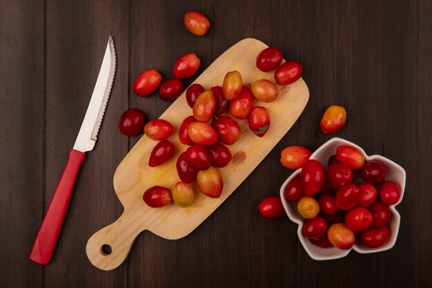 Vue de dessus des cerises de cornaline acidulée sur une planche de cuisine en bois avec un couteau avec des cerises de cornaline sur un bol sur un mur en bois