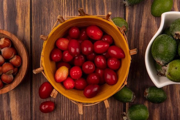 Vue de dessus des cerises de cornaline sucrées sur un seau avec des noisettes sur un bol en bois avec feijoas sur un bol sur un mur en bois