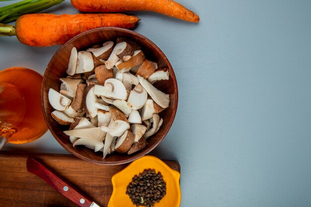 Vue de dessus de champignons frais tranchés dans un bol et un couteau de cuisine avec des grains de poivre noir sur une planche à découper en bois et des carottes fraîches sur bleu clair avec copie espace