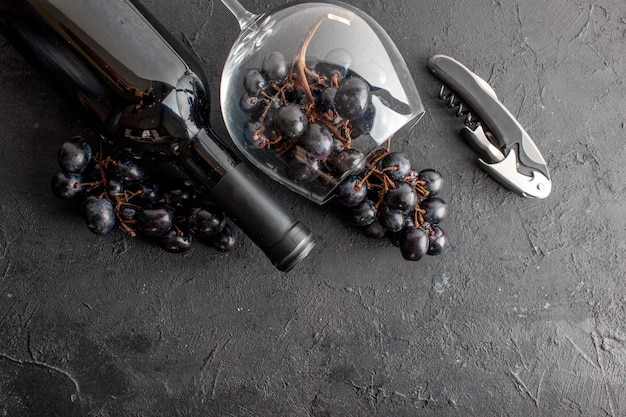 Photo gratuite vue de dessus de charmants raisins noirs dans un verre à vin renversé et un ouvre-bouteille de vin sur un sol sombre