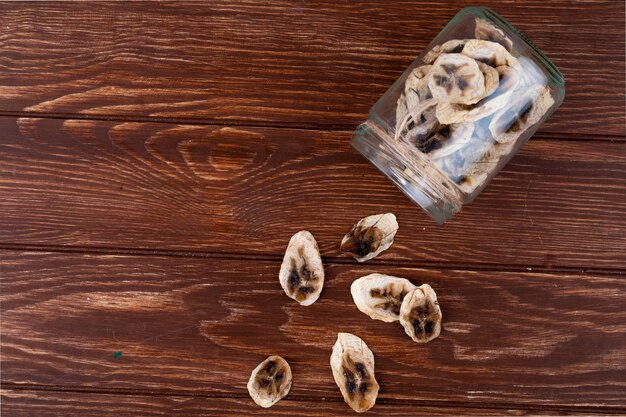 Vue de dessus des chips de banane séchées éparpillées dans un bocal en verre sur fond de bois avec copie espace